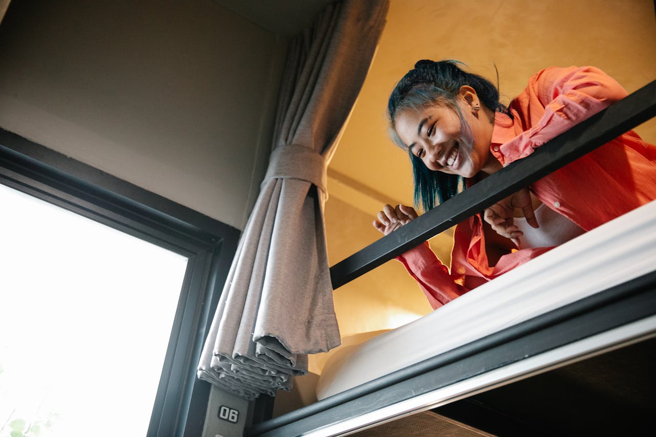 Home Low angle of young ethnic cheerful female resting on bunk bed and looking down from top tier of bed near window with curtain